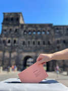 Ein Umschlag wird in eine Urne gesteckt, im Hintergrund sieht man verschwommen die Porta Nigra
