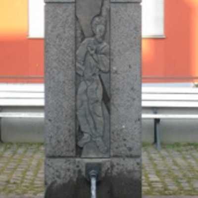 Brunnenstock aus Basalt mit einem Bronzerelief des Heiligen Andreas mit seinem typischen Attribut, dem Kreuz.